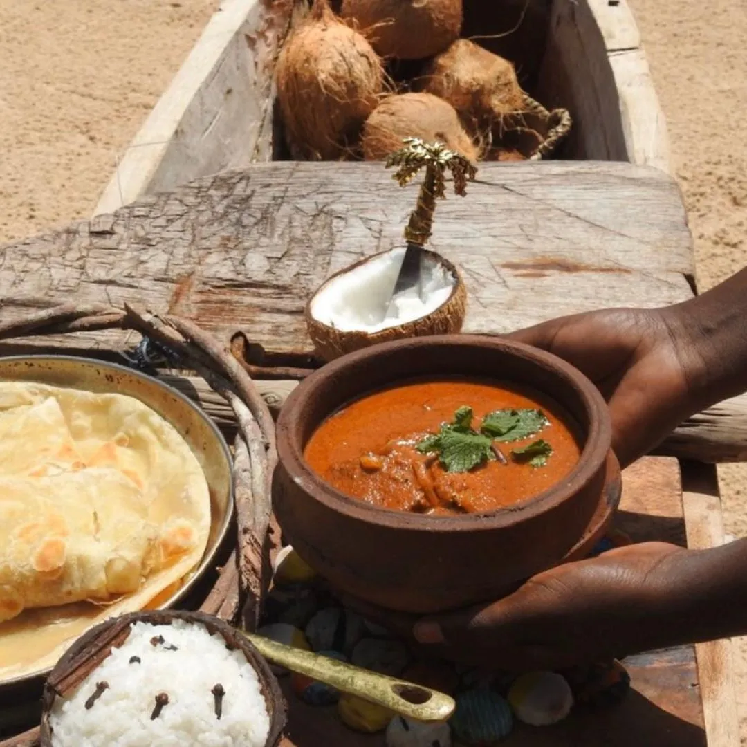 Hands holding a clay pot of rich orange curry topped with fresh mint, served alongside flatbread, spiced rice in a coconut shell, and whole coconuts on a rustic wooden table.