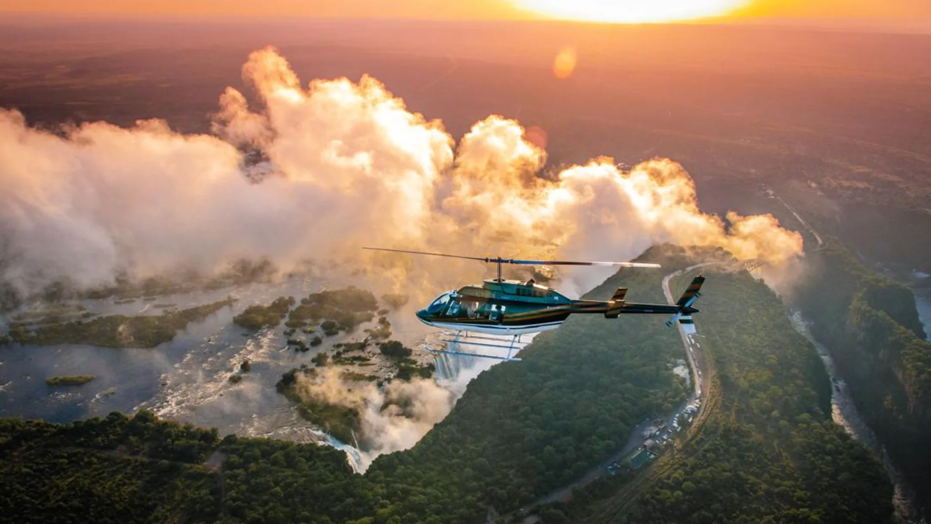 Aerial view of a helicopter flying over a massive waterfall surrounded by lush green forests during a golden sunset, with mist rising high into the sky