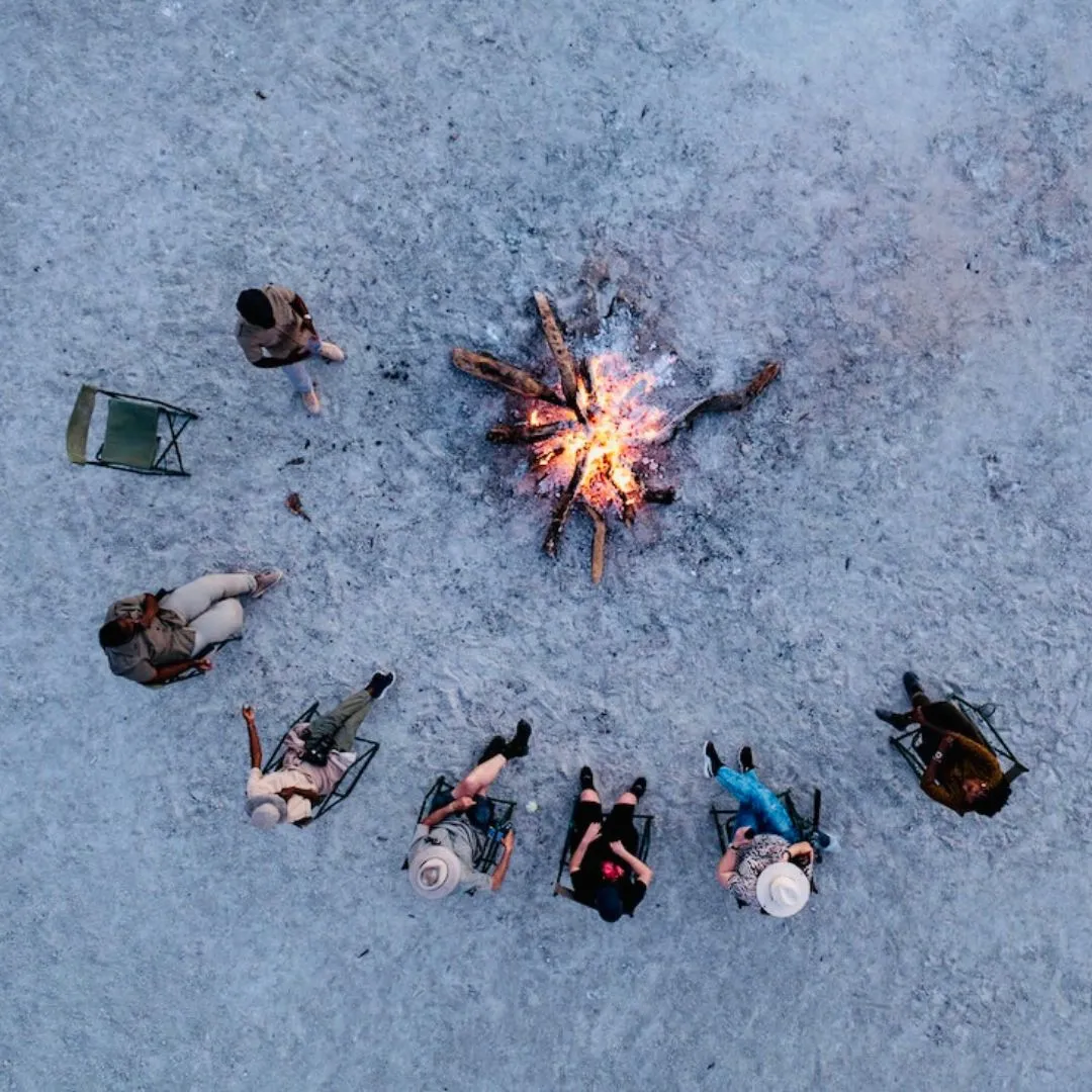 Top-down aerial view of a group of people sitting in lawn chairs in a semi-circle around a blazing campfire on a pale, sandy surface.