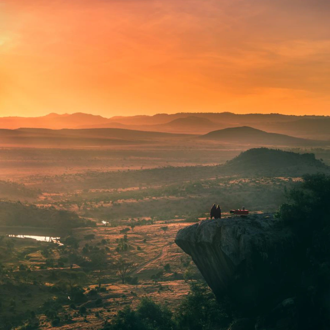 Two people standing on a rocky cliff overlook, watching a vibrant orange sunset over a vast valley landscape with rolling hills